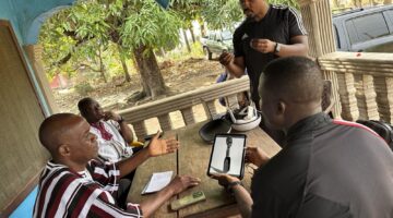 On a shaded porch in Sanniquellie, four collaborators gather around a table as one displays an archival photo of a carved spoon on a tablet; a VR headset and laptop sit nearby.