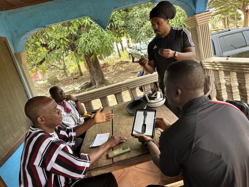 On a shaded porch in Sanniquellie, four collaborators gather around a table as one displays an archival photo of a carved spoon on a tablet; a VR headset and laptop sit nearby.